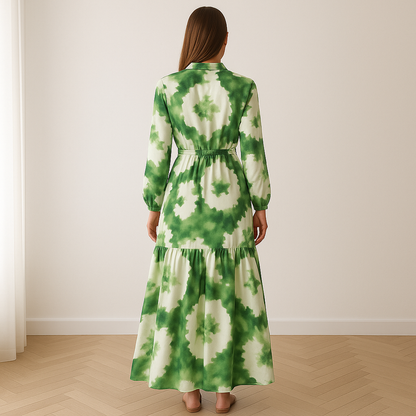 Vrouw in een elegante, lange maxi-jurk met groen-wit bloemenpatroon, gemaakt van luchtige stof. De jurk heeft lange mouwen, een strikceintuur in de taille en een vloeiende rok zonder naad onder de knie. Beschikbaar in maten S t/m 2XL.