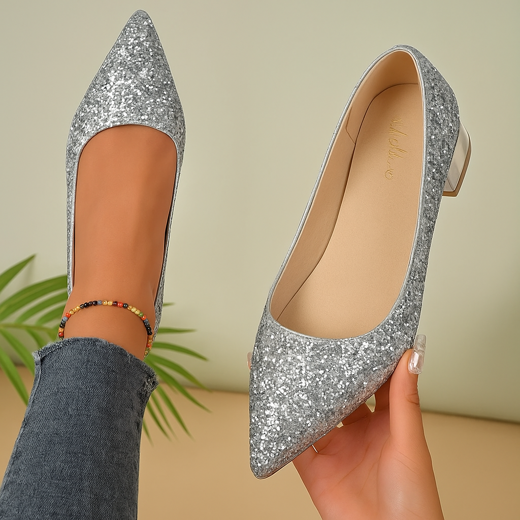 Zilverkleurige glitterballerina's voor dames met een chroomkleurige hak en spitse neus, gedragen met een denim jeans. Eén schoen wordt vastgehouden om de zachte beige binnenzool met gouden logo te tonen. Perfect voor feestelijke of chique gelegenheden.